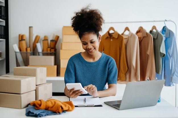 Shopify Development 1 A smiling small business owner in a clothing workshop using a calculator and laptop to manage sales and inventory, surrounded by stacked shipping boxes and a garment rack.