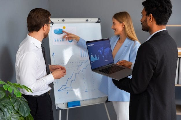 Three business professionals discussing ERP strategy; a consultant points to growth charts on a whiteboard while another holds a laptop displaying real-time data analytics.