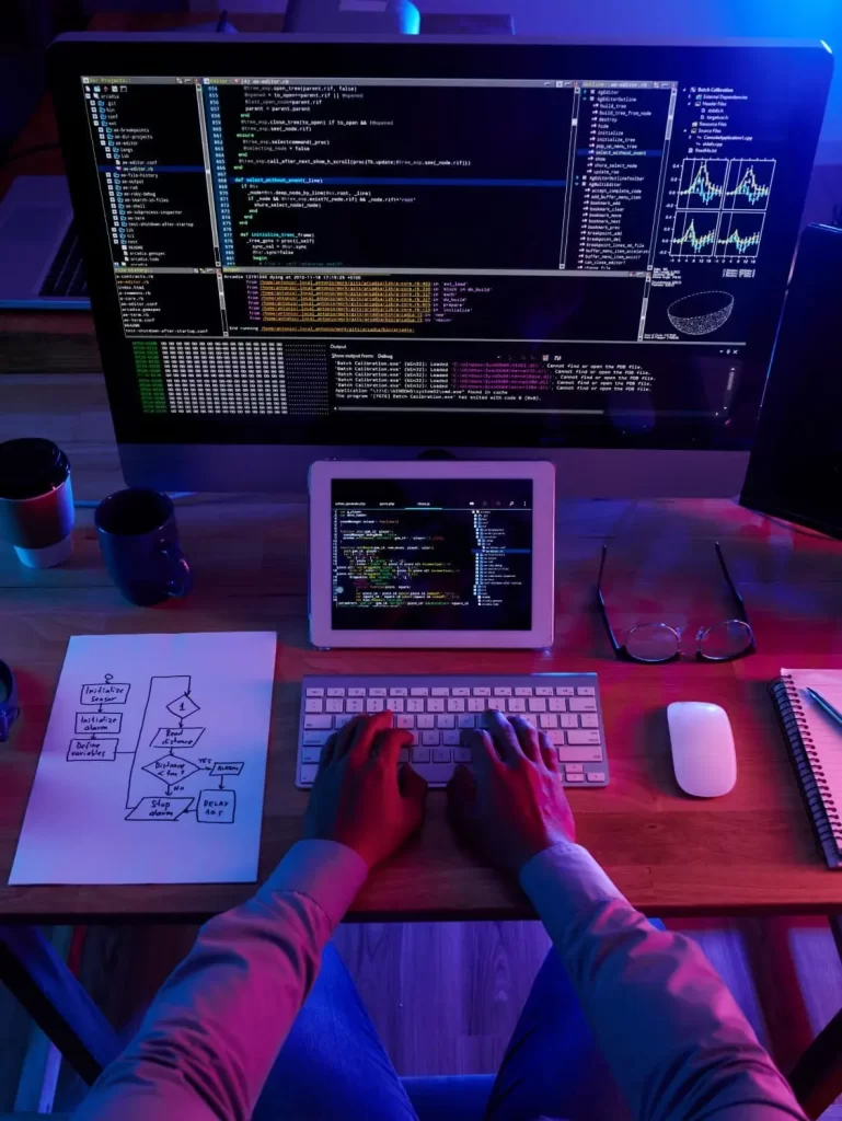 eCommerce 2 High-angle view of a developer's hands typing on a wireless keyboard at a wooden desk illuminated by blue and purple lights, featuring a large monitor displaying code, a tablet, and a handwritten logic flowchart on paper.